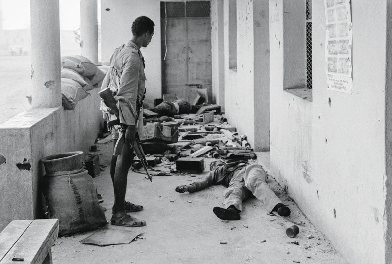 Banco de Etiopía, recién terminado el asalto. Los cadáveres de dos de sus defensores, observados por uno de los atacantes. Al que yace en primer plano ya le han quitado las botas y cuantas armas y objetos de valor que llevaba encima. Un momento después se hará saltar la caja fuerte del interior con dinamita y veré amontonarse sobre una mesa de billar los fajos de billetes y las monedas de oro. Es la primera vez en mi vida –también la última– que participo en el atraco a un banco.