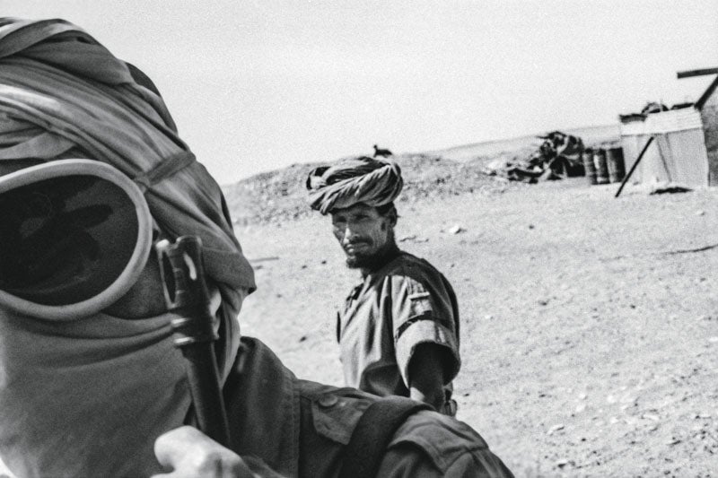 Le tengo especial afecto a esta fotografía, que nunca vi positivada hasta que Jeosm se encargó de ello. El hombre del turbante que mira a la cámara es el ex cabo de la Policía Territorial española Belali uld Mahrabi. Belali era muy amigo mío, y tras haber vivido aventuras juntos antes de la Marcha Verde, lo encontré un año más tarde en la guerrilla del Frente Polisario, combatiendo contra Marruecos. Días después de tomar esta imagen desde el vehículo que me iba a transportar a otro lugar del Sáhara, Belali murió combatiendo en Uad Ashram. Cayó herido y quedó atrás, me contaron, y mientras sus compañeros se retiraban estuvieron oyendo los disparos de su Kalashnikov hasta que agotó la munición y llegó el silencio.