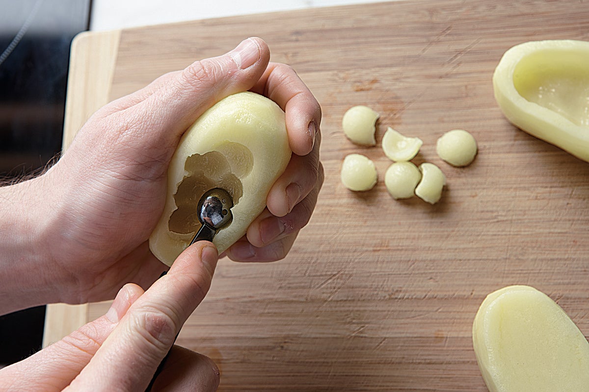 Para empezar con la receta de patatas rellenas de manitas de cerdo, pelamos y tallamos las patatas en forma ovalada y, con un sacabolas, les hacemos un hueco en el medio.