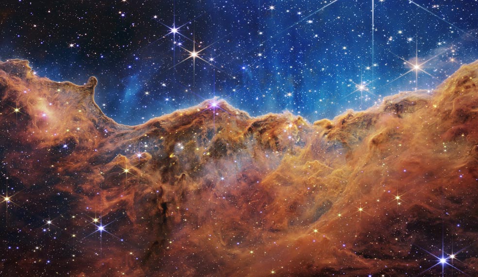 Este paisaje de 'montañas' y 'valles' salpicados de estrellas brillantes es el borde de una región cercana y joven donde se forman estrellas, en la nebulosa de Carina. La imagen, capturada en luz infrarroja por telescopio espacial James Webb, revela por primera vez regiones de nacimiento estelar que antes eran invisibles.